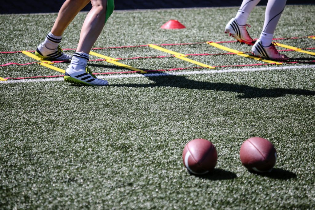 Football players practice agility drills on a sunlit grass field with ladders and balls.